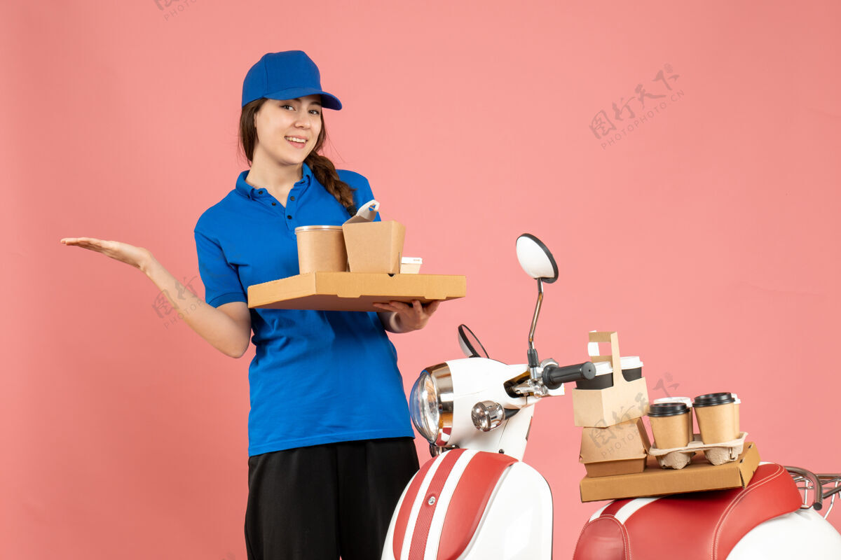 蛋糕自信的女快递员站在摩托车旁 手拿咖啡和小蛋糕 背景是柔和的桃色粉彩成人咖啡