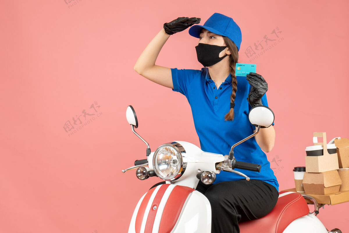 银行前视图集中快递女孩戴着医用面罩和手套坐在滑板车上拿着银行卡在粉彩桃色背景下传递订单坐摩托车前面