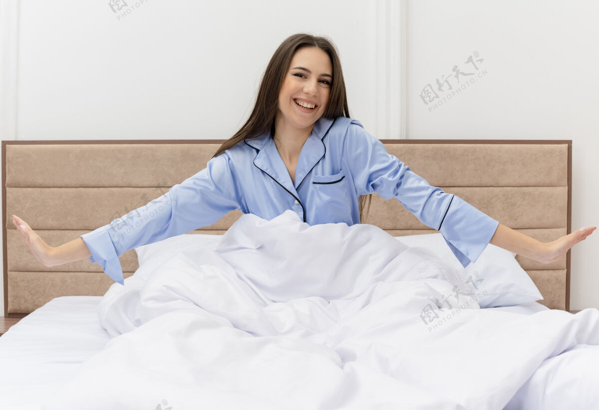 背景穿着蓝色睡衣的年轻美女坐在床上 枕在柔软的枕头上 快乐而积极地微笑着醒来 在卧室里享受周末卧室享受灯光