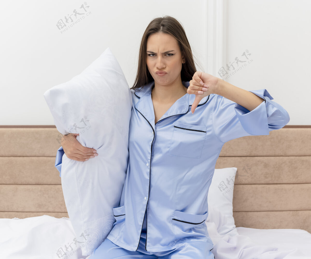 床穿着蓝色睡衣的年轻美女坐在床上 枕头不高兴地在卧室里竖起大拇指相机不高兴女人