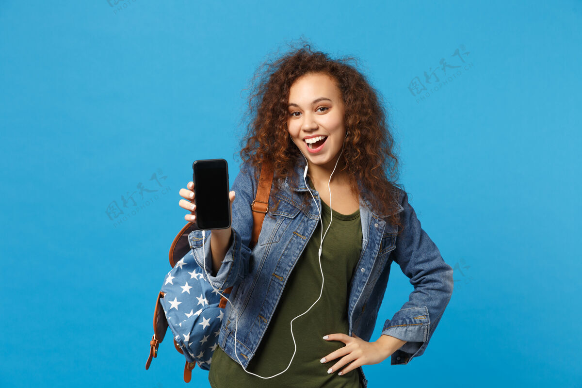 耳机年轻的非洲裔美国少女 穿着牛仔服 背着耳机 隔离在蓝色的墙上屏幕青少年人物