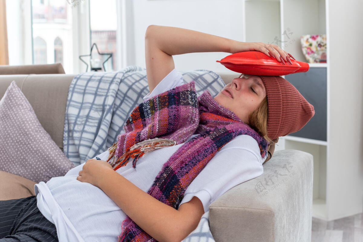 年轻不健康的年轻女子戴着帽子 脖子上围着暖和的围巾 感觉不舒服 生病 患感冒和流感 头上戴着热水瓶 躺在客厅的沙发上 看起来很担心感觉周围生病