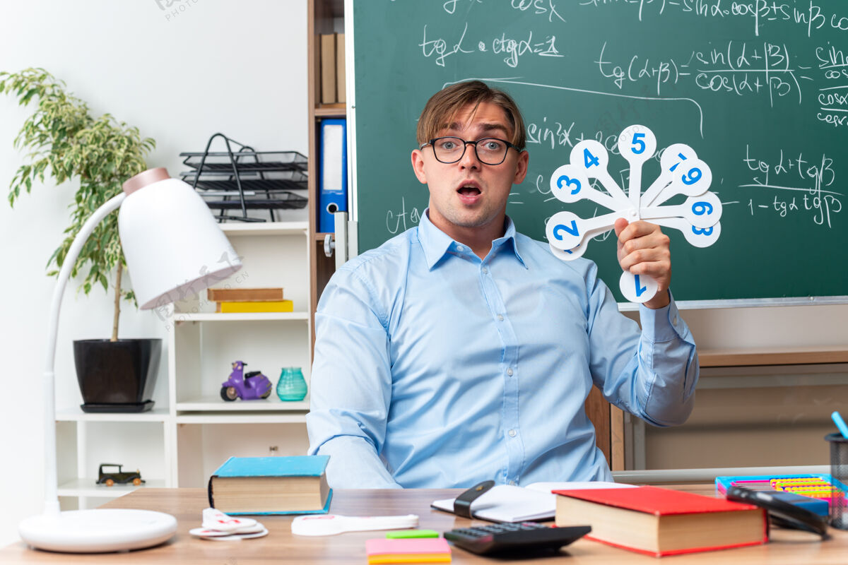 教室年轻的男老师戴着眼镜 展示着解释课的号牌 惊讶地坐在课桌旁 教室的黑板前放着书和笔记课笔记学校