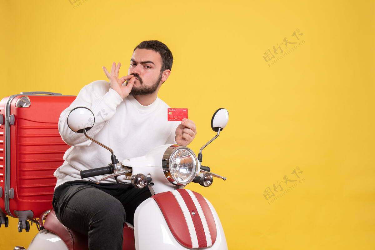 车辆年轻自信的旅游者坐在摩托车上 提着手提箱 手里拿着银行卡 在黄色上做着完美的手势体育摩托车旅游
