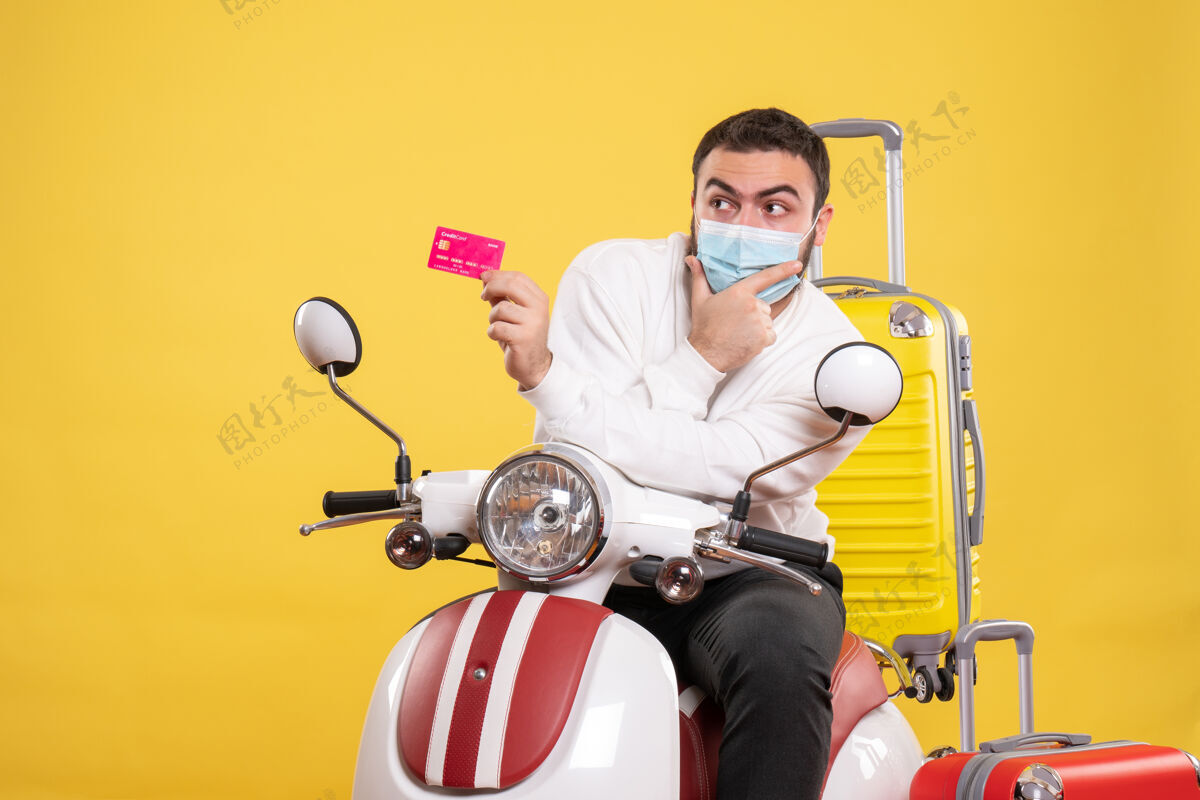 坐旅行概念的俯视图 戴着医用面罩的年轻人坐在摩托车上 黄色手提箱 手里拿着银行卡高尔夫银行球员