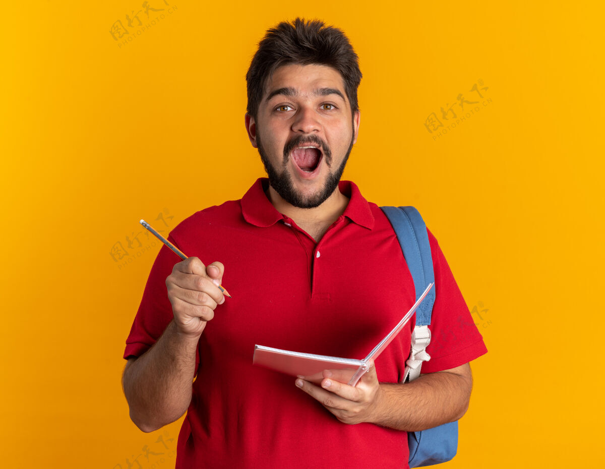 姿势年轻的留着胡子的学生 穿着红色马球衫 背着背包 手里拿着笔记本和铅笔 站在橙色的墙上 惊讶地发现了新的想法站胡须惊喜