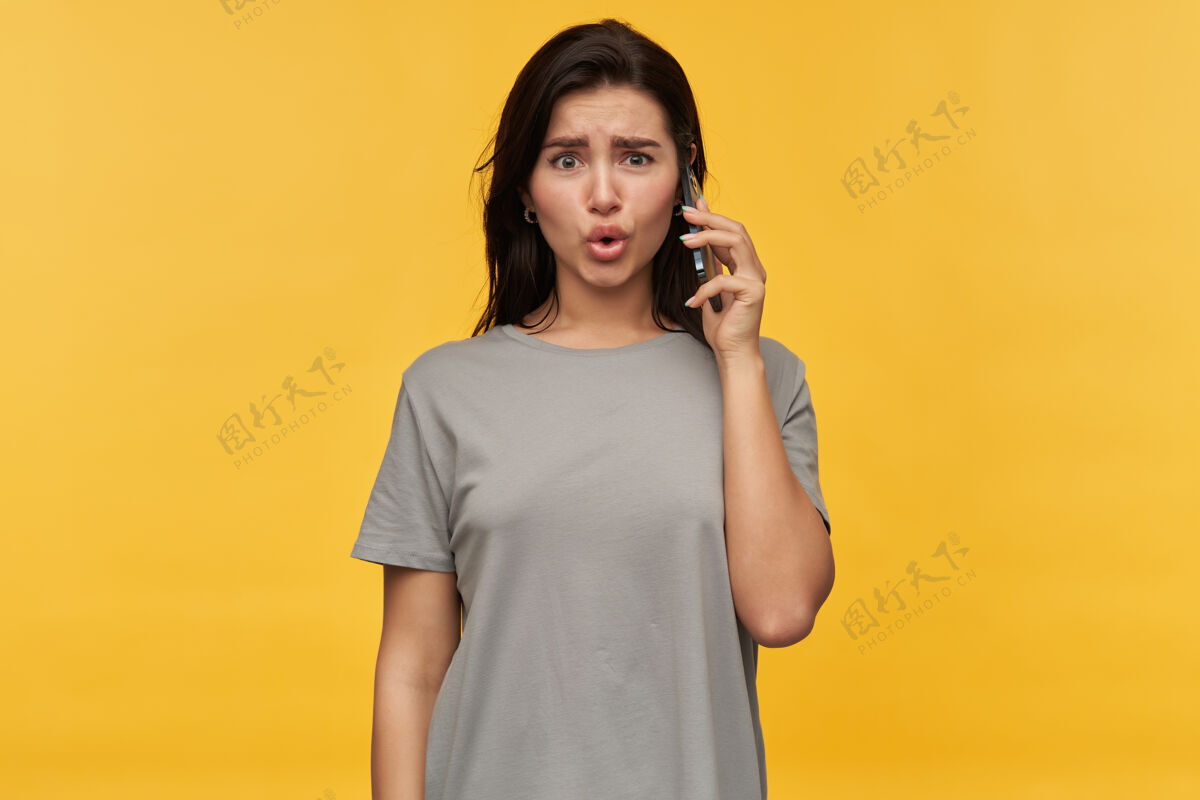 肖像一个身穿灰色T恤的黑发年轻女子在黄色的墙上打电话 这让她大吃一惊黑发站着可爱