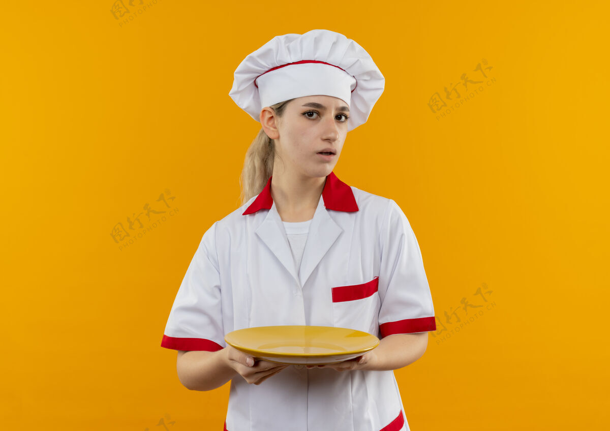 漂亮年轻漂亮的厨师穿着厨师制服 拿着空盘子 孤立地站在橙色的墙上盘子制服厨师