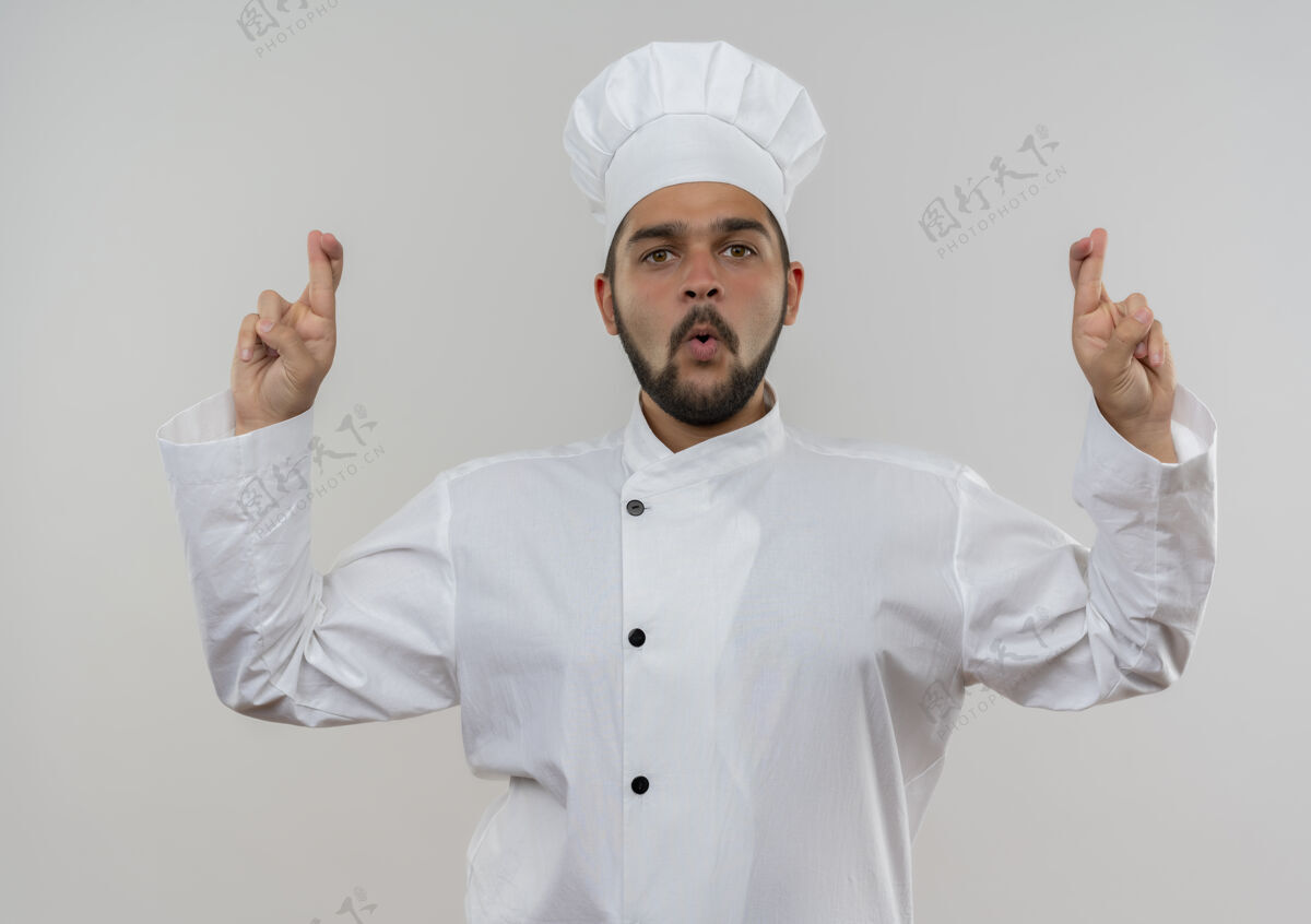 十字令人印象深刻的年轻男厨师在厨师制服做交叉手指手势隔离在白墙上厨师男性制服