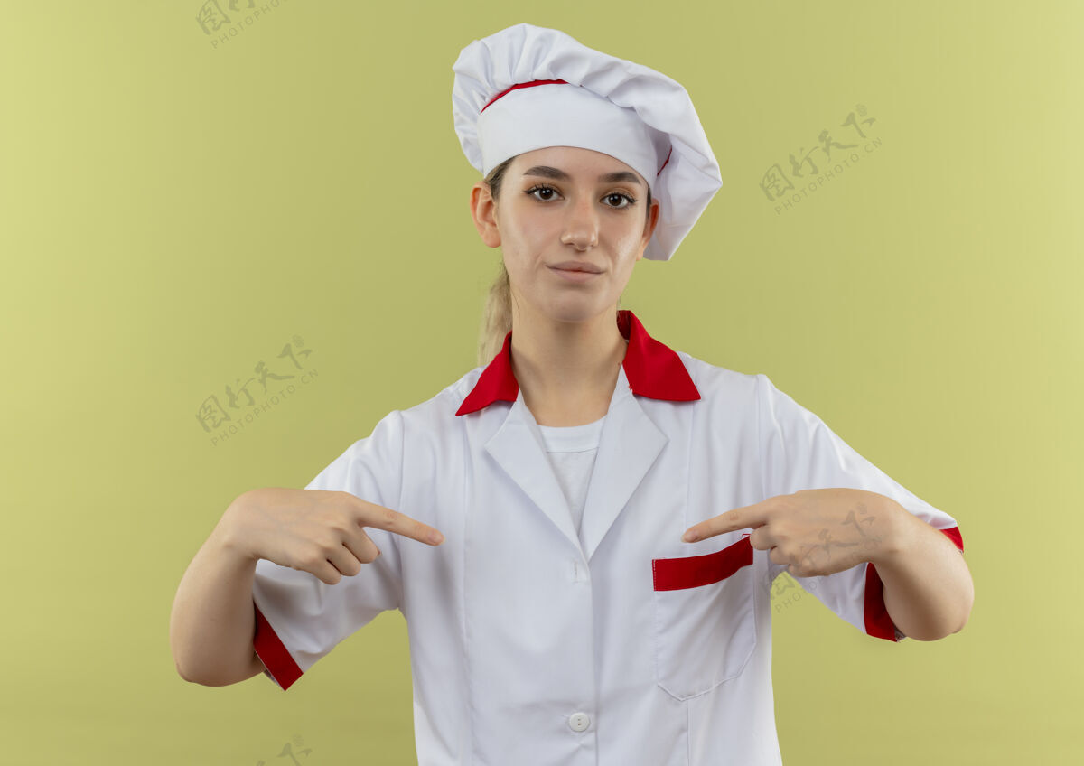 她自己穿着厨师制服的自信年轻漂亮的厨师指着绿色墙壁上孤立的自己漂亮烹饪自信