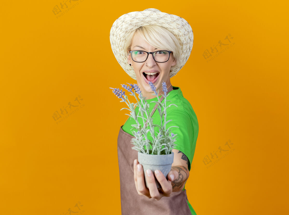 站一位年轻的园丁 围裙短发 戴着帽子 站在橙色的背景下 脸上挂着盆栽植物的微笑锅头发微笑