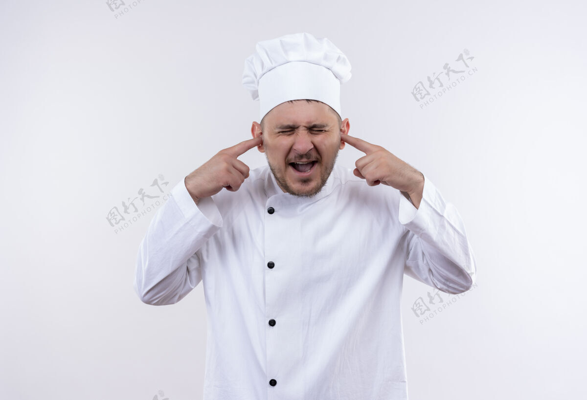 年轻穿着厨师制服的年轻帅哥生气地把手指放进耳朵里 闭着眼睛孤立在白墙上恼火厨师封闭