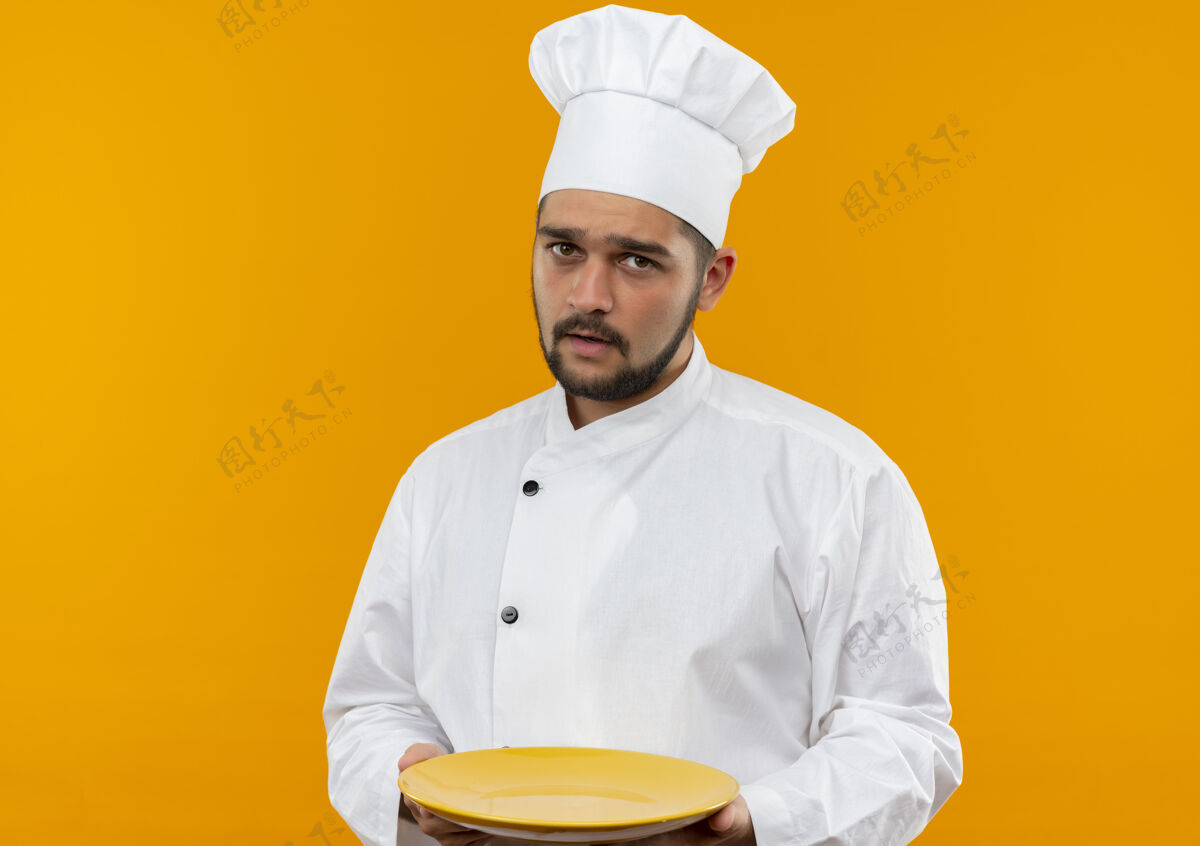 烹饪令人印象深刻的年轻男厨师在厨师制服举行空盘子隔离在橙色墙上空制服年轻