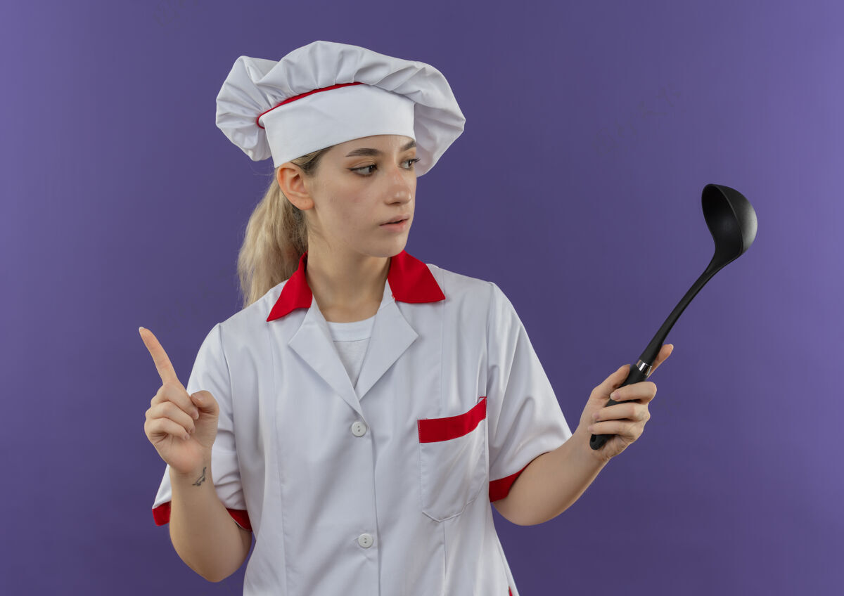 烹饪令人印象深刻的年轻漂亮厨师在厨师制服举行 并看着勺子和提高手指孤立在紫色墙上手指制服手持