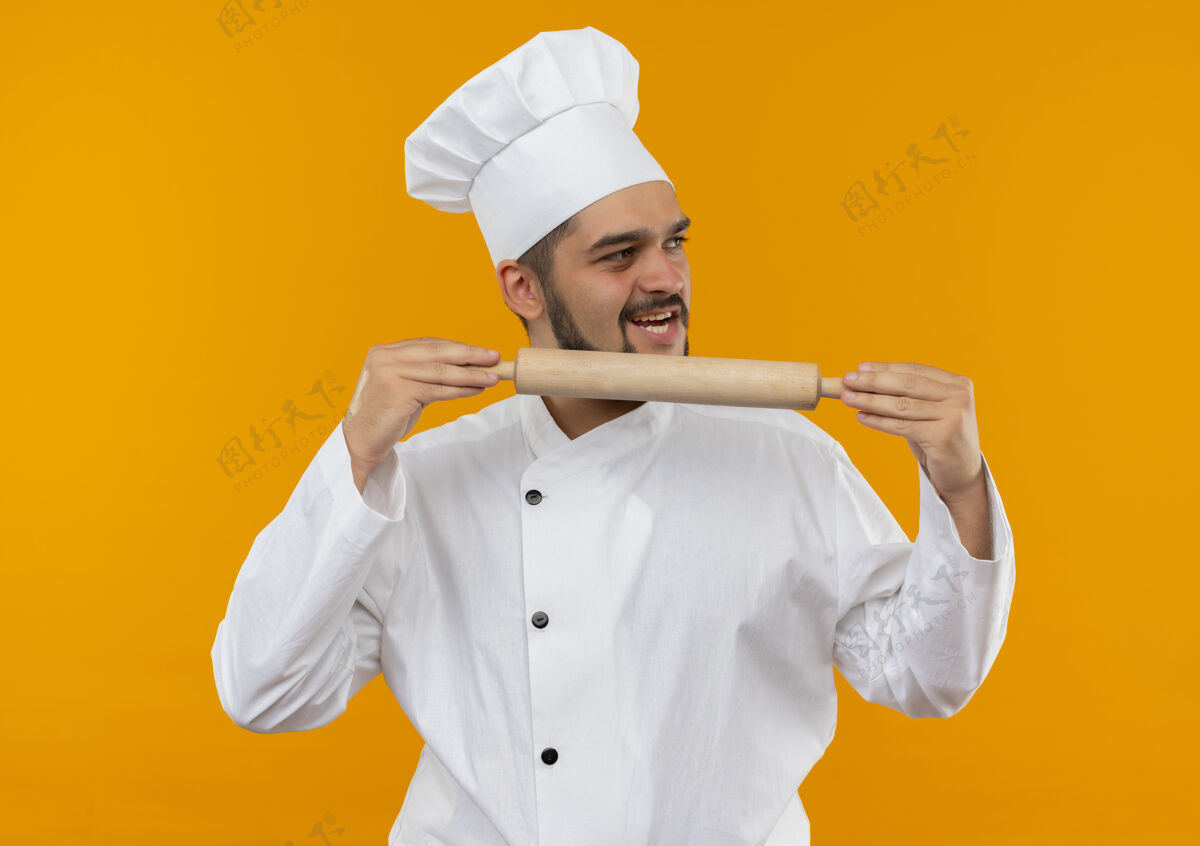 抱快乐的年轻男厨师身着厨师制服 手持擀面杖 看着橙色墙上孤立的一面男欢乐卷