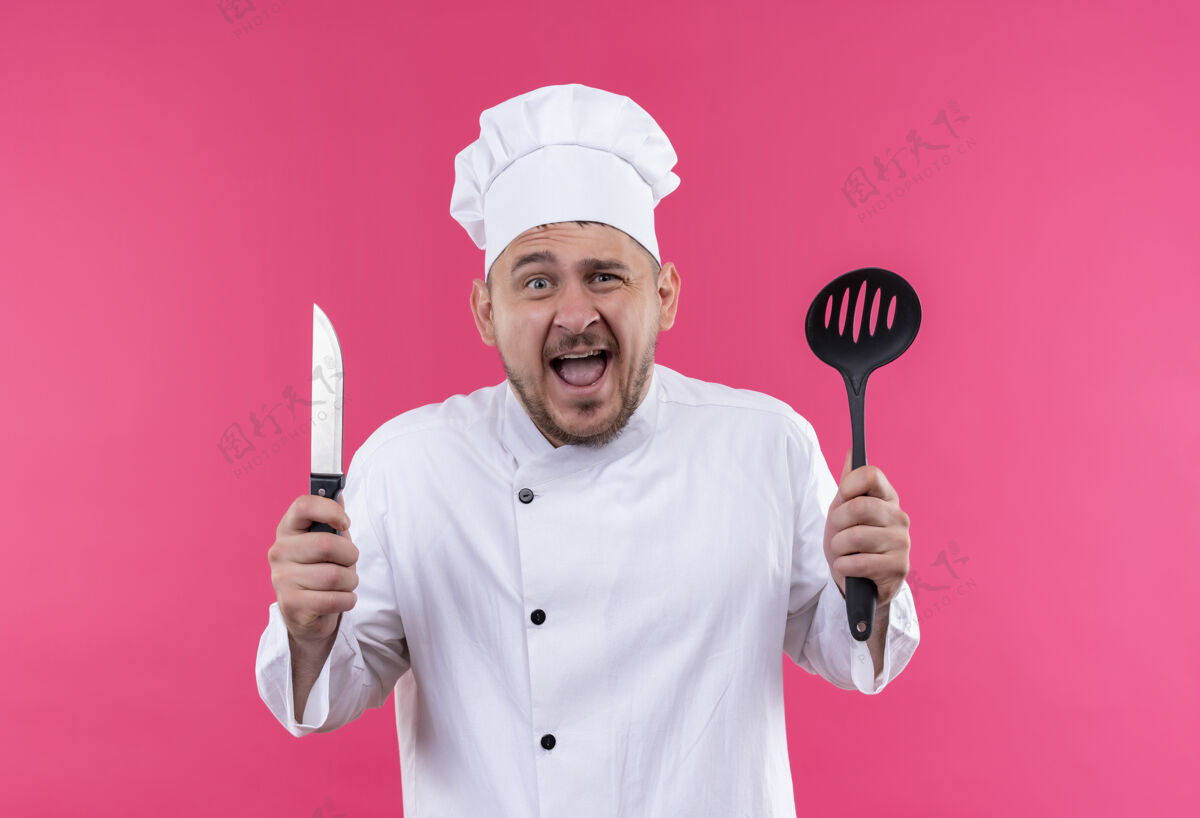 印象深刻年轻帅气的厨师穿着厨师制服 手里拿着开槽汤匙和刀 隔离在粉红色的墙上手持厨师帅气