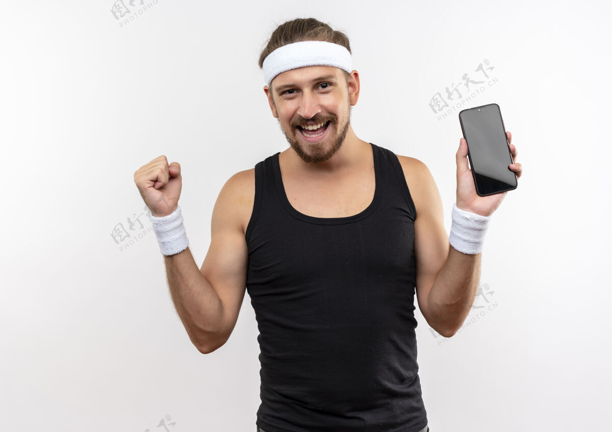 男人快乐的年轻帅气的运动男戴着头带和腕带拿着手机紧握拳头孤立在白墙上手机手机握紧