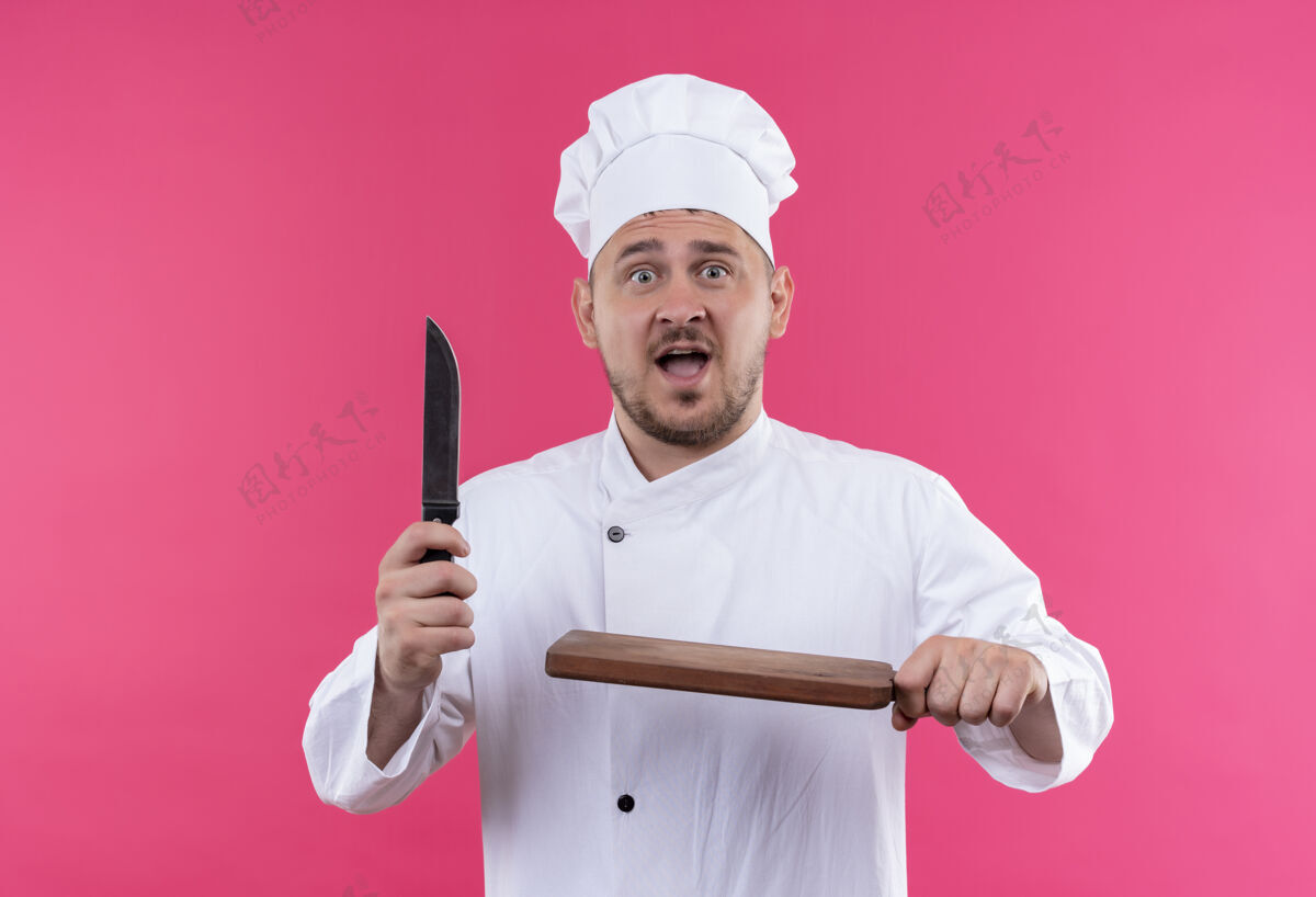 刀年轻帅气的厨师穿着厨师制服 手里拿着刀和砧板 隔离在粉红色的墙上持有板削减