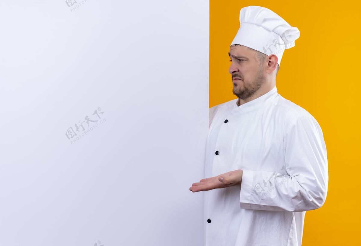 年轻困惑的年轻帅哥厨师穿着厨师制服站在白色的墙后 用手指着隔离在橙色墙上的复制空间困惑指向制服