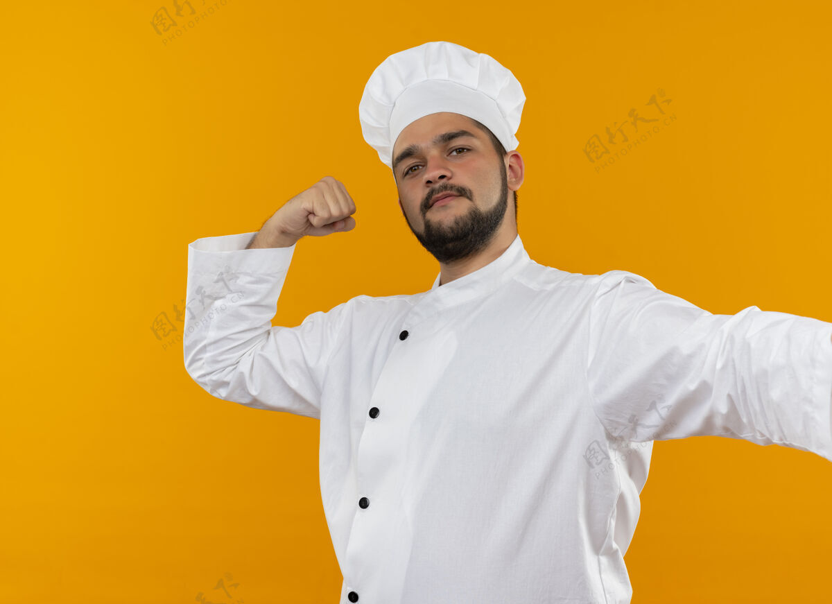 姿态自信的年轻男厨师身着厨师制服 在橙色的墙上做着坚强的手势厨师强壮制服