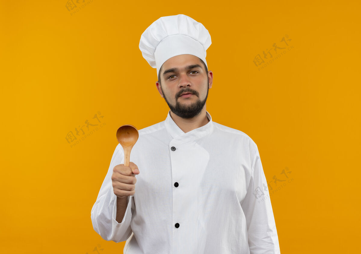 持有自信的年轻男性厨师身着厨师制服 拿着勺子 隔离在橙色的墙上 留有复印空间年轻烹饪男性