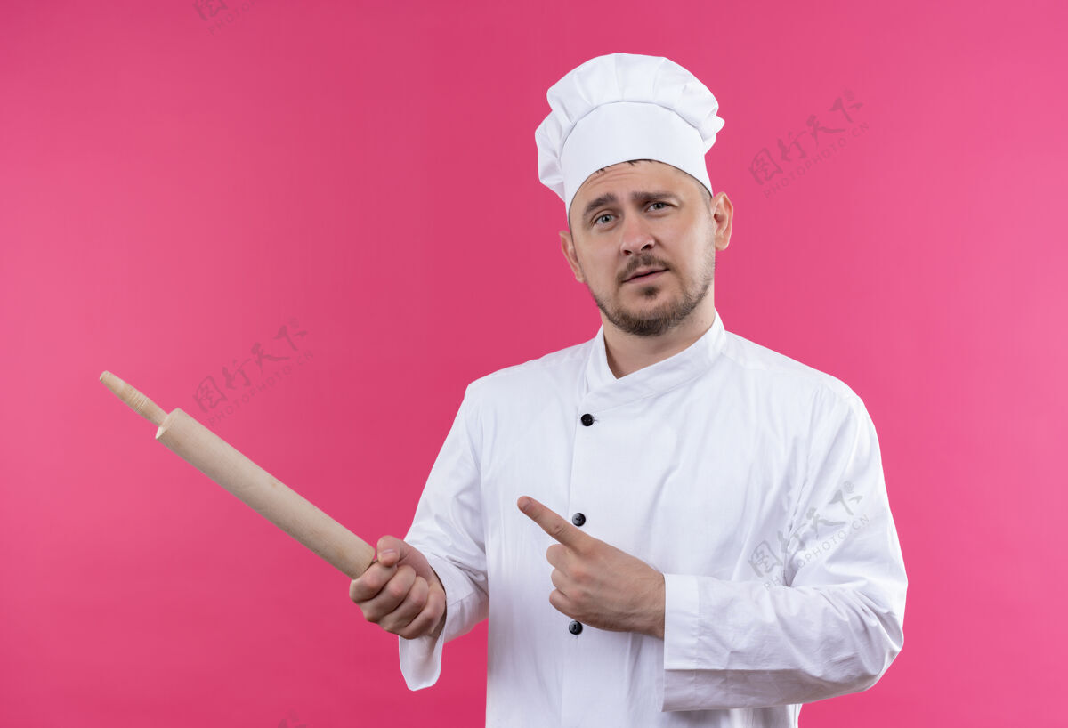 指着自信的年轻帅哥厨师穿着厨师制服拿着擀面杖指着粉色墙上孤立的一面拿着自信制服