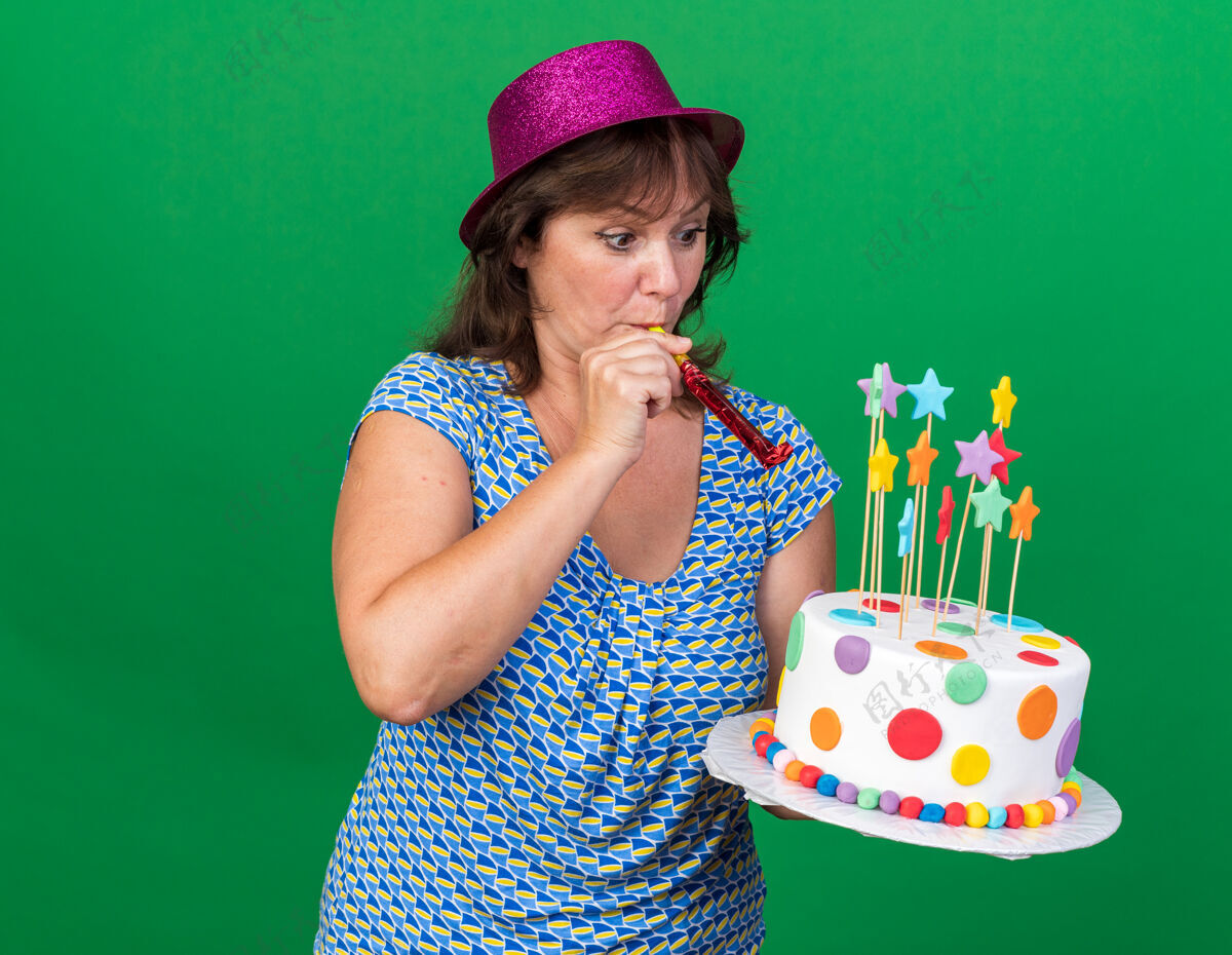 中间中年妇女戴着生日帽 手持生日蛋糕吹着口哨 站在绿色的墙壁上庆祝生日蛋糕举行哨子