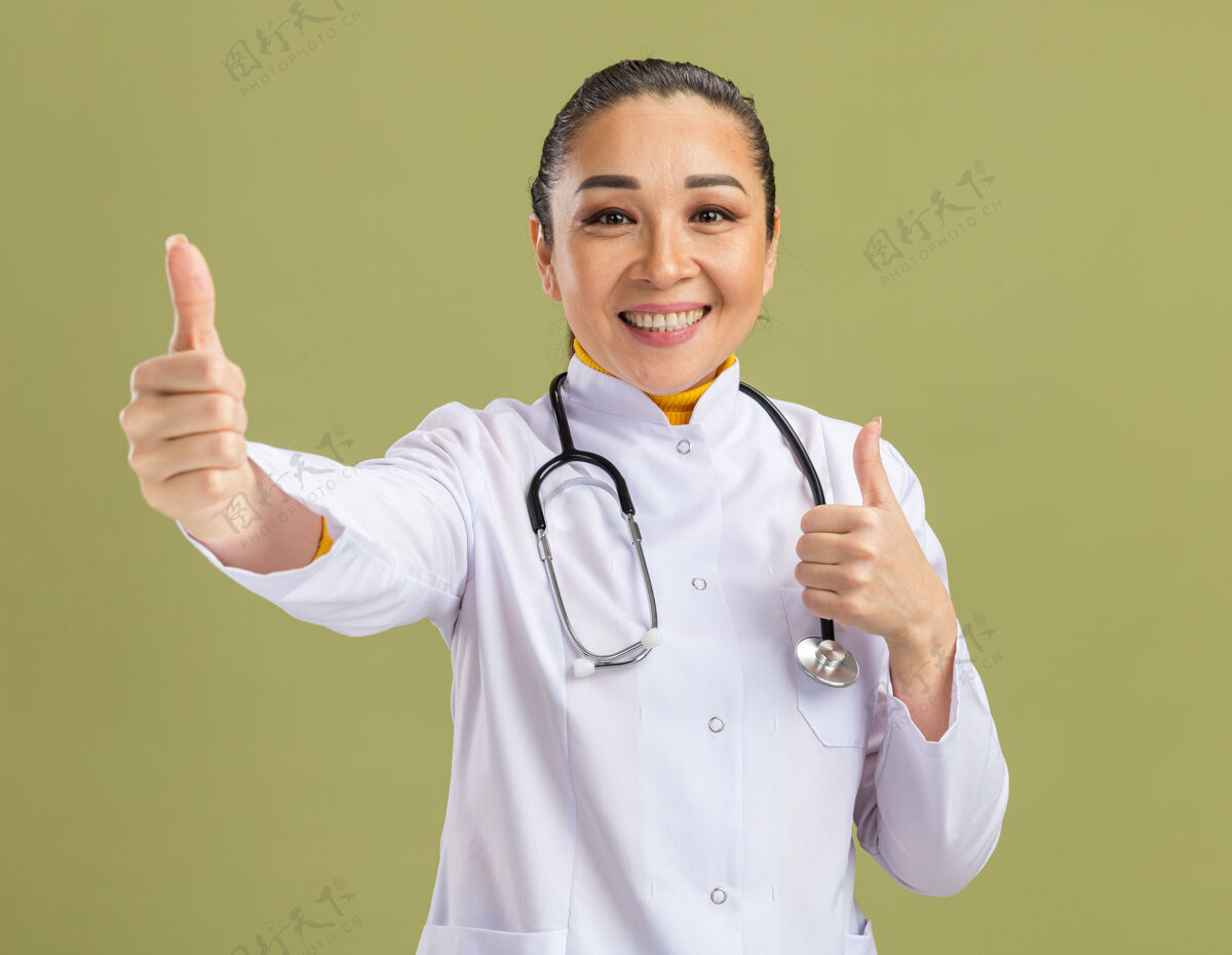年轻人身穿白色药衣 脖子上戴着听诊器的年轻女医生微笑着自信地竖起大拇指站在绿色的墙上听诊器脖子周围
