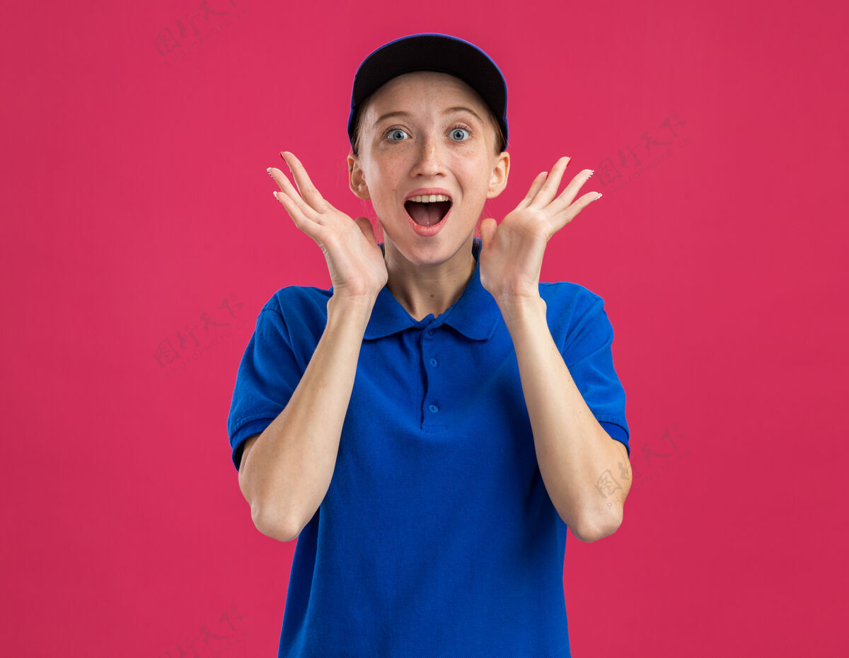 举起身穿蓝色制服 头戴鸭舌帽的年轻送货员高兴又惊讶地举起双臂站在粉红色的墙上接生帽子手臂