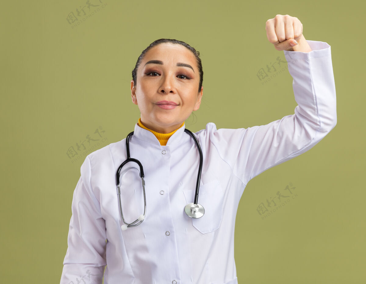 站身穿白色药衣的年轻女医生脖子上戴着听诊器握紧拳头站在绿色的墙上 快乐而自信外套握紧听诊器