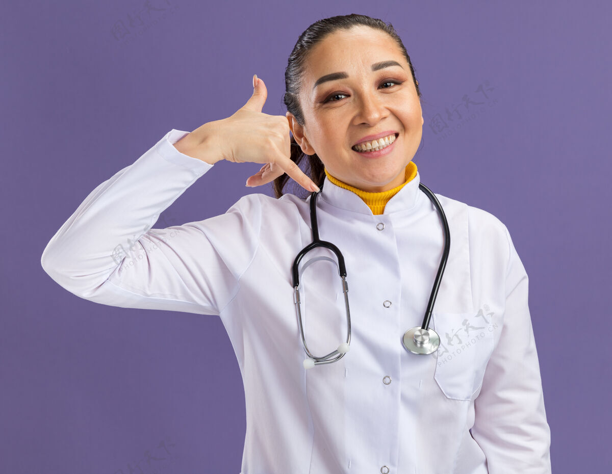 外套年轻的女医生穿着白药衣 脖子上戴着听诊器 脸上带着微笑 站在紫色的墙上做着叫我的手势医生听诊器手势