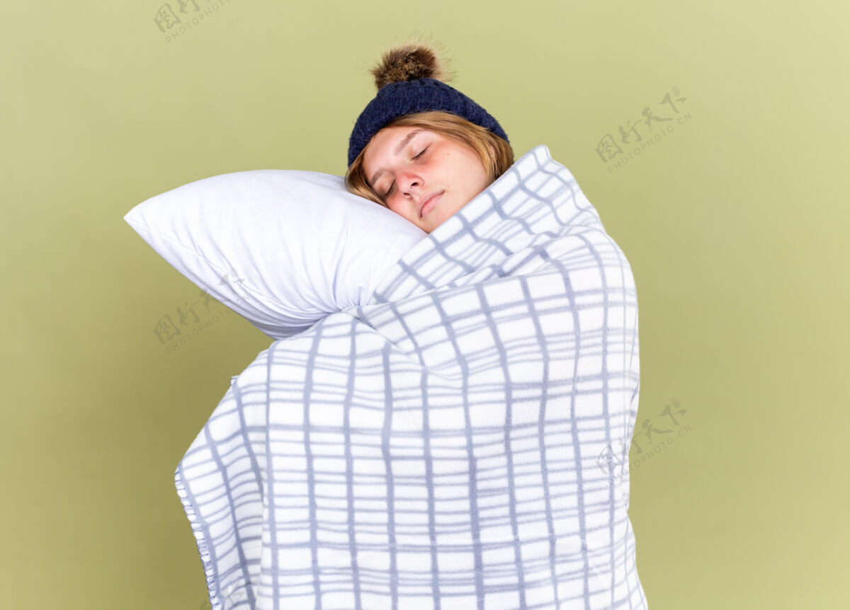温暖不健康的年轻女子戴着暖和的帽子 裹着毯子 抱着枕头 闭着眼睛 站在绿色的墙上睡觉女人戴着枕头