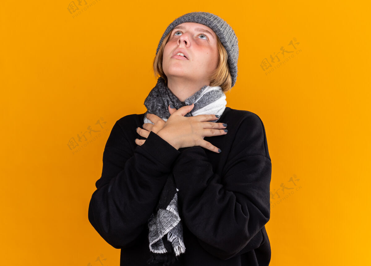 希望不健康的年轻女子戴着暖和的帽子 脖子上围着围巾 感觉不舒服 患了感冒和流感 站在橘色的墙上仰望着希望的表情痛苦感冒表情