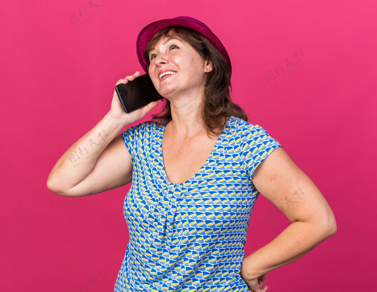 庆祝戴着派对帽的中年妇女一边打电话一边开心地笑着 站在粉色的墙上庆祝生日派对微笑年龄站立