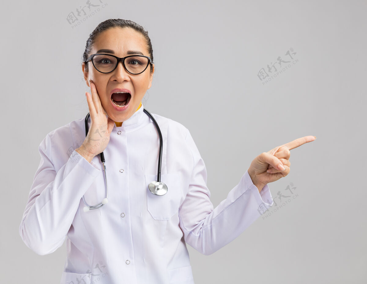 外套年轻的女医生穿着白色医用外套 脖子上戴着听诊器 惊讶地用食指指着站在白墙上的一边脖子听诊器女人