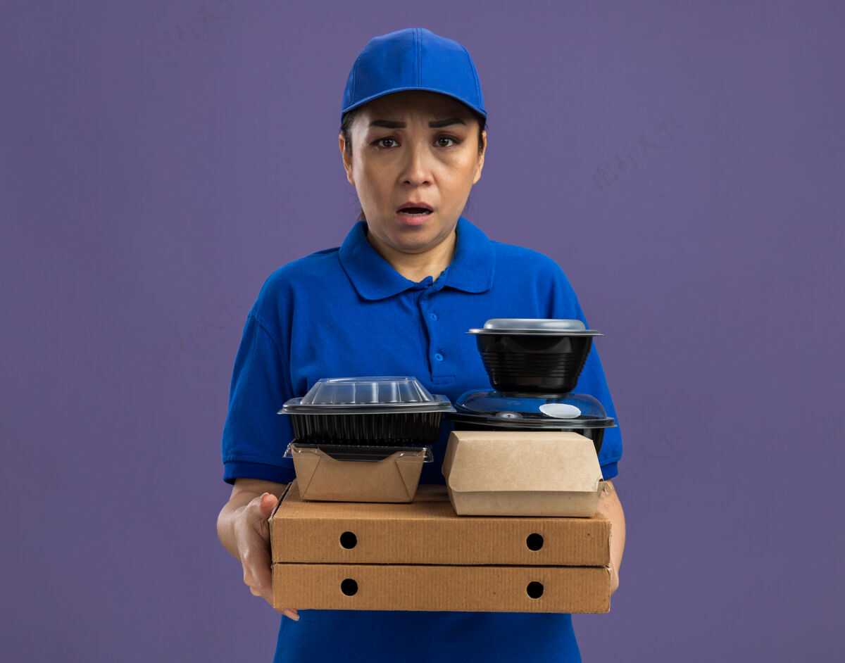 女人身穿蓝色制服 头戴鸭舌帽的年轻女送货员站在紫色的墙上 手里拿着披萨盒和食品包 严肃地皱着眉头拿着食物盒子