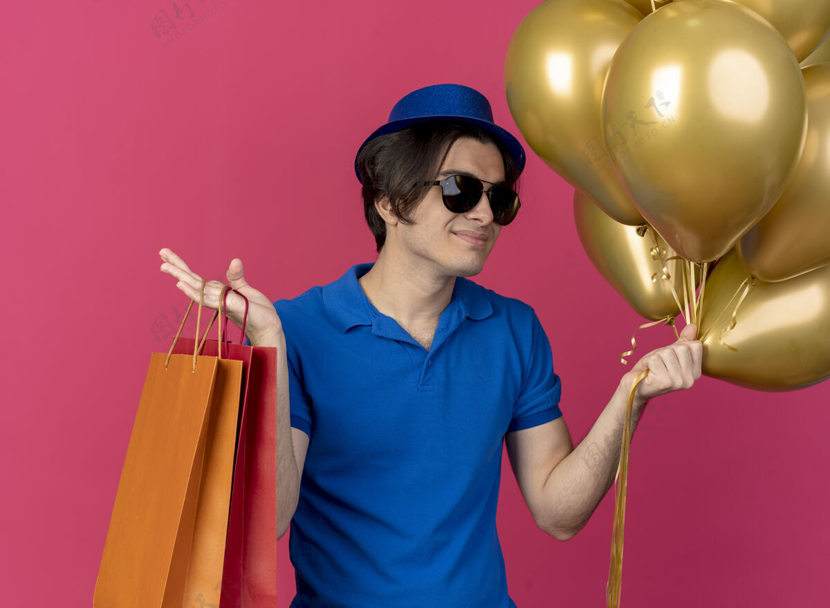 自信自信英俊的白种人戴着太阳眼镜 戴着蓝色派对帽 手里拿着氦气球和纸购物袋空间男人气球
