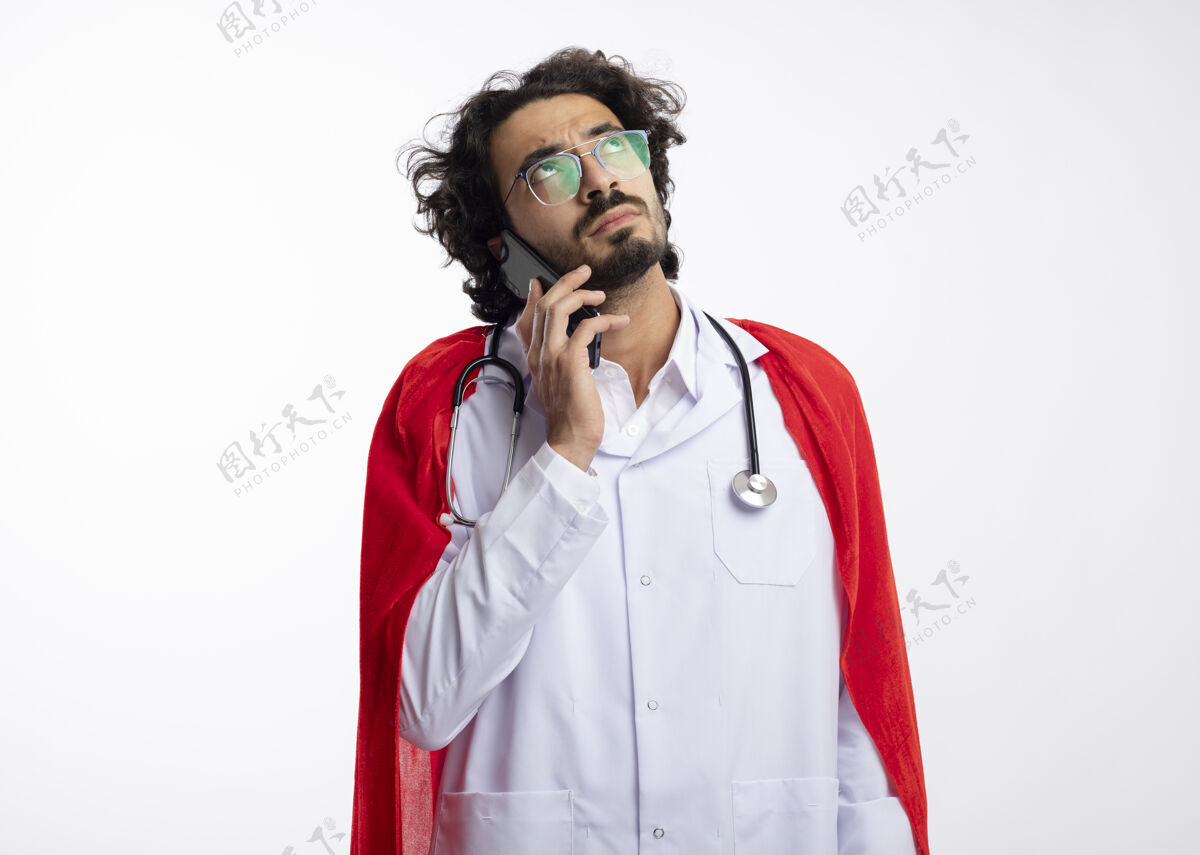 周围自信的年轻白种人超级英雄戴着眼镜 身穿医生制服 披着红色斗篷 脖子上戴着听诊器 在电话里交谈斗篷光学穿
