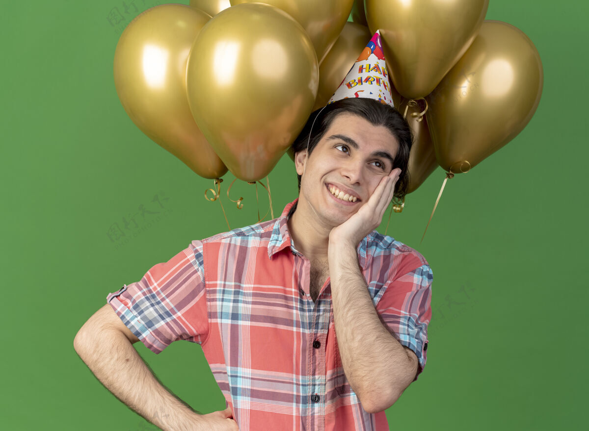 脸高兴的高加索帅哥戴着生日帽站在氦气球前把手放在脸上穿帽子氦