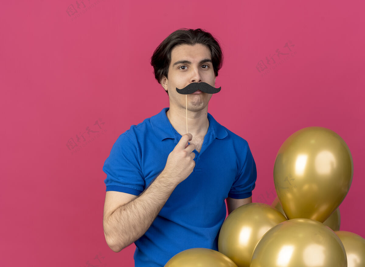假的滑稽英俊的白人男子戴着蓝色的派对帽 手里拿着氦气球和假胡子粉色搞笑派对