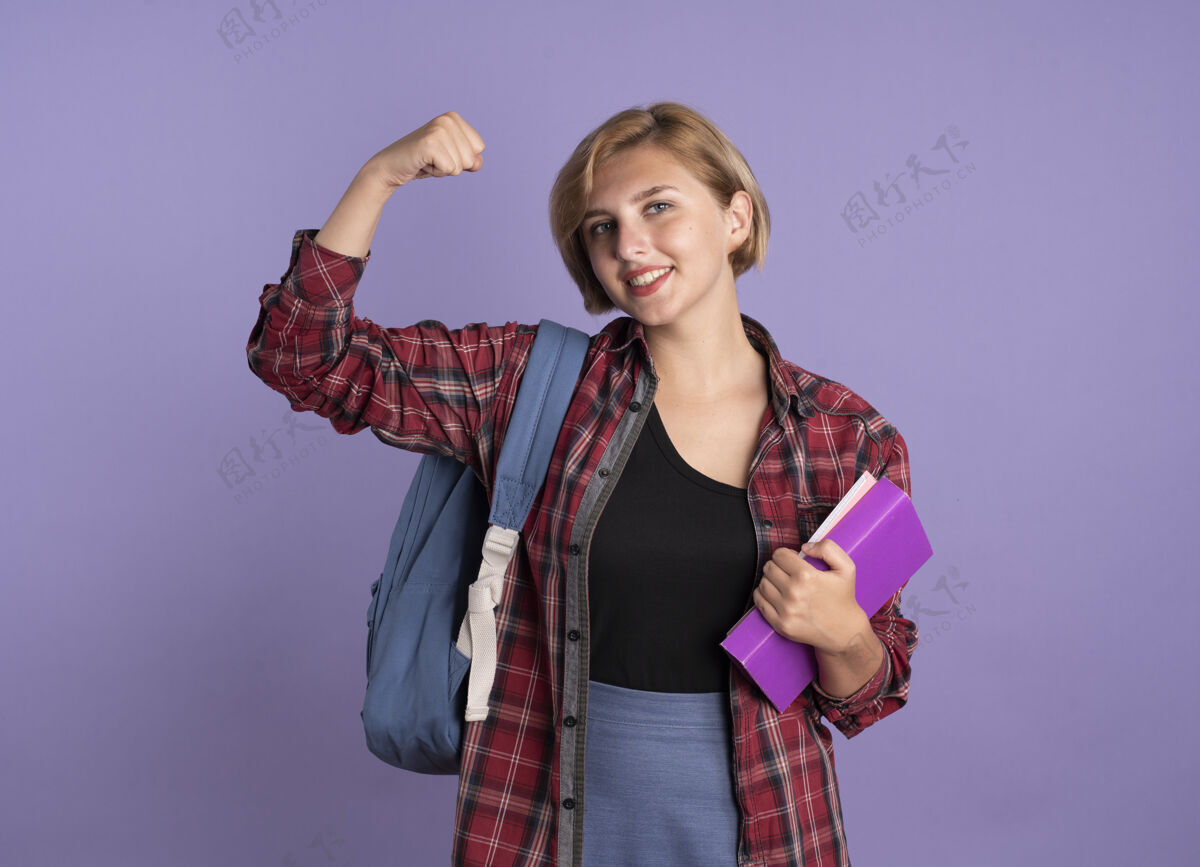 微笑微笑的年轻斯拉夫学生女孩穿着背包时态二头肌拿着书和笔记本年轻紫色女孩