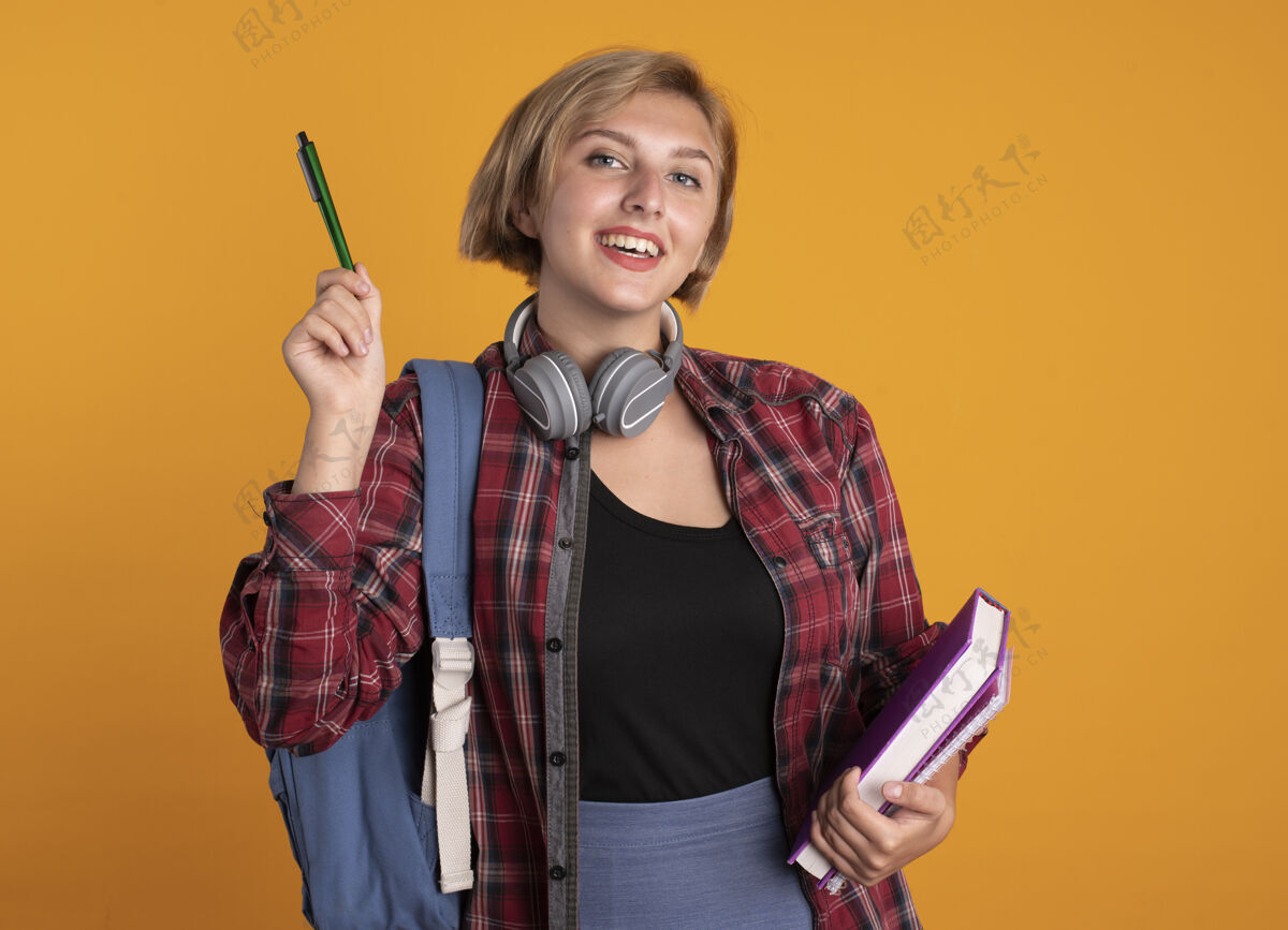 背包微笑的年轻斯拉夫女学生戴着耳机 背着背包 手里拿着笔本和笔记本斯拉夫人穿钢笔