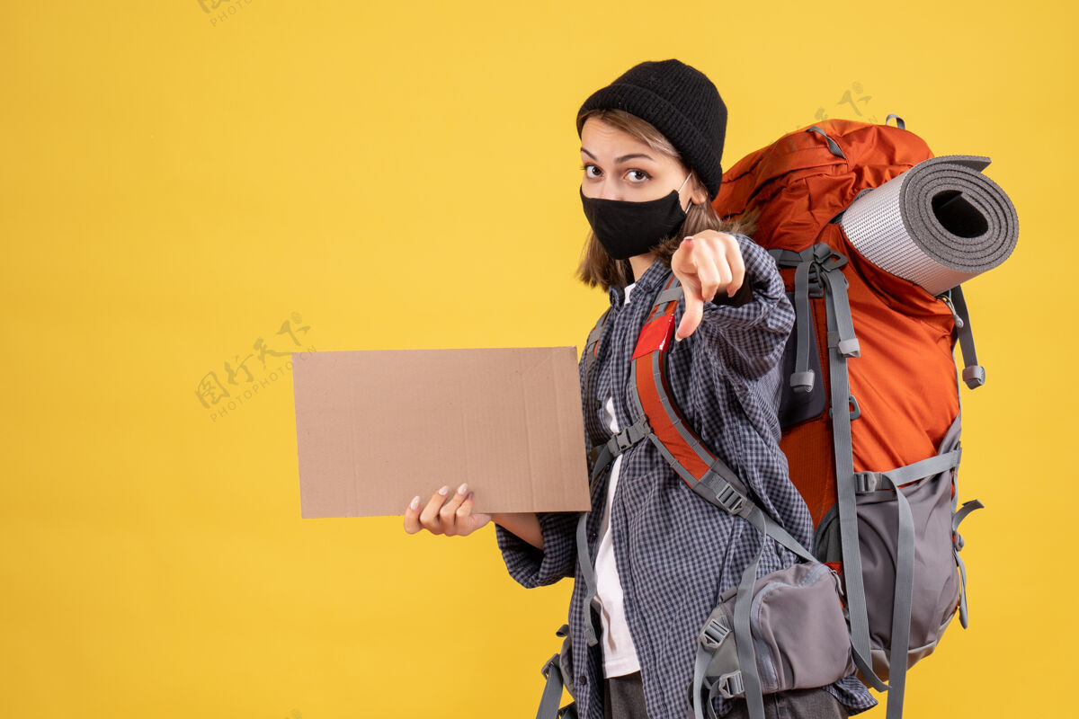 肖像带着黑色面具和背包的旅行女孩拿着纸板指着摄像机成人面具指点