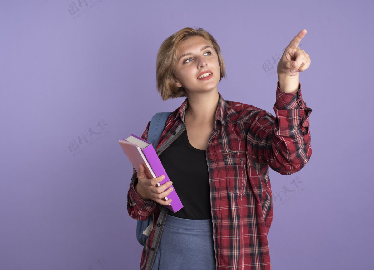 微笑带着微笑的年轻斯拉夫学生女孩背着书包拿着书和笔记本看着并指着旁边壁板年轻笔记本