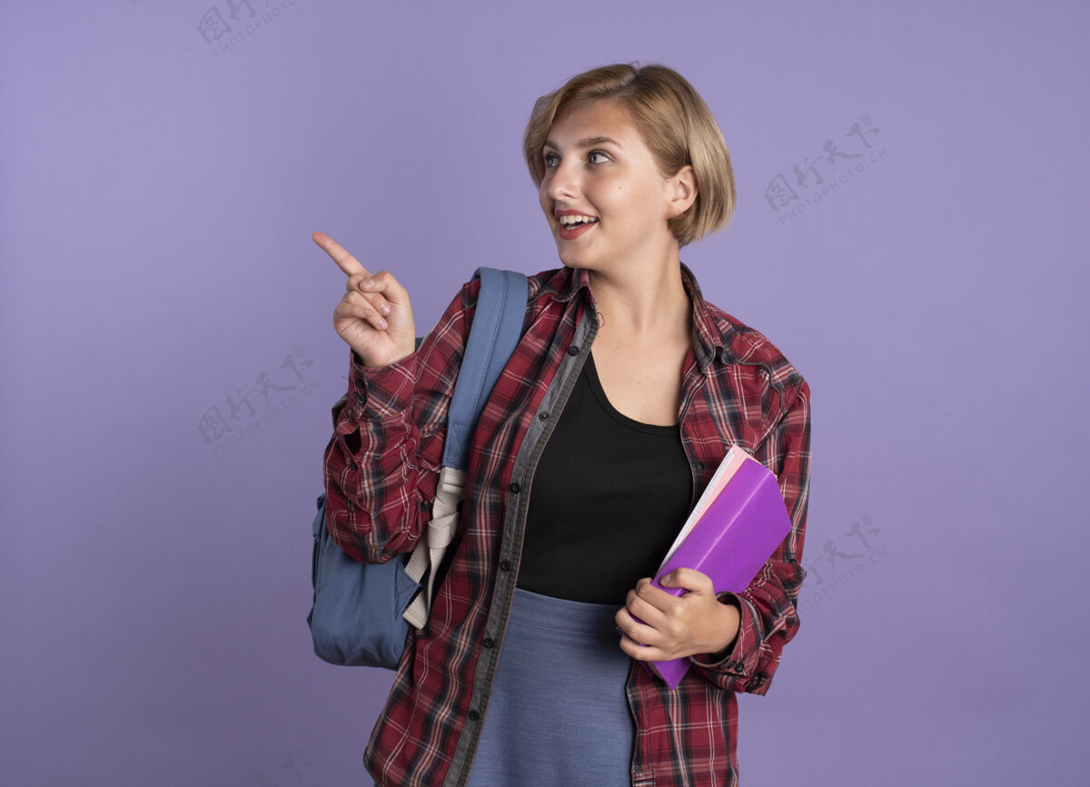 紫色惊讶的年轻斯拉夫学生女孩背着书包拿着书和笔记本看着并指着旁边穿复制指向