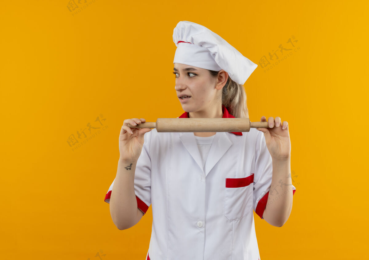 年轻年轻漂亮的厨师穿着厨师制服 手持擀面杖 看着橙色墙壁上孤立的一面 留着复印空间卷壁板烹饪