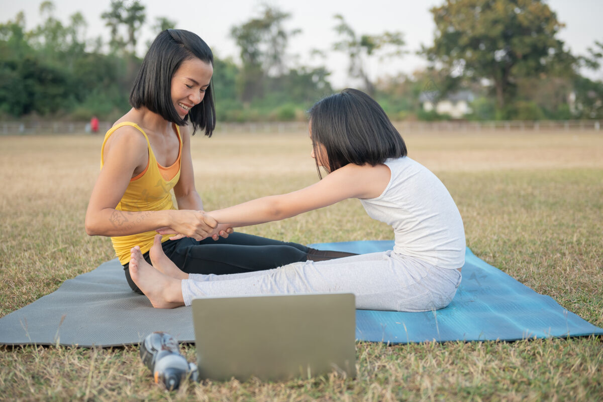 瑜伽妈妈和女儿在做瑜伽在公园里训练妇女和孩子户外运动健康的运动生活方式 在线观看瑜伽练习视频教程和坐姿前弯姿势健康视频笔记本电脑