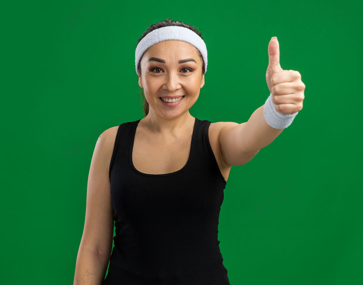 脸戴着头巾和臂章 脸上带着微笑 竖起大拇指的年轻健身女士站在绿色的墙上头带健康秀