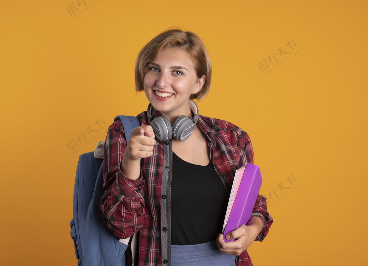 学生微笑着的年轻斯拉夫女学生戴着耳机 背着背包 拿着书和笔记本点着相机微笑斯拉夫笔记本