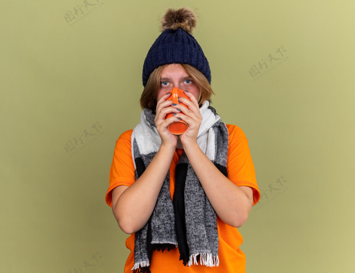 热不健康的年轻女子戴着帽子 脖子上围着围巾 感觉不舒服 喝着热茶 站在绿色的墙壁上患感冒和流感穿茶流感